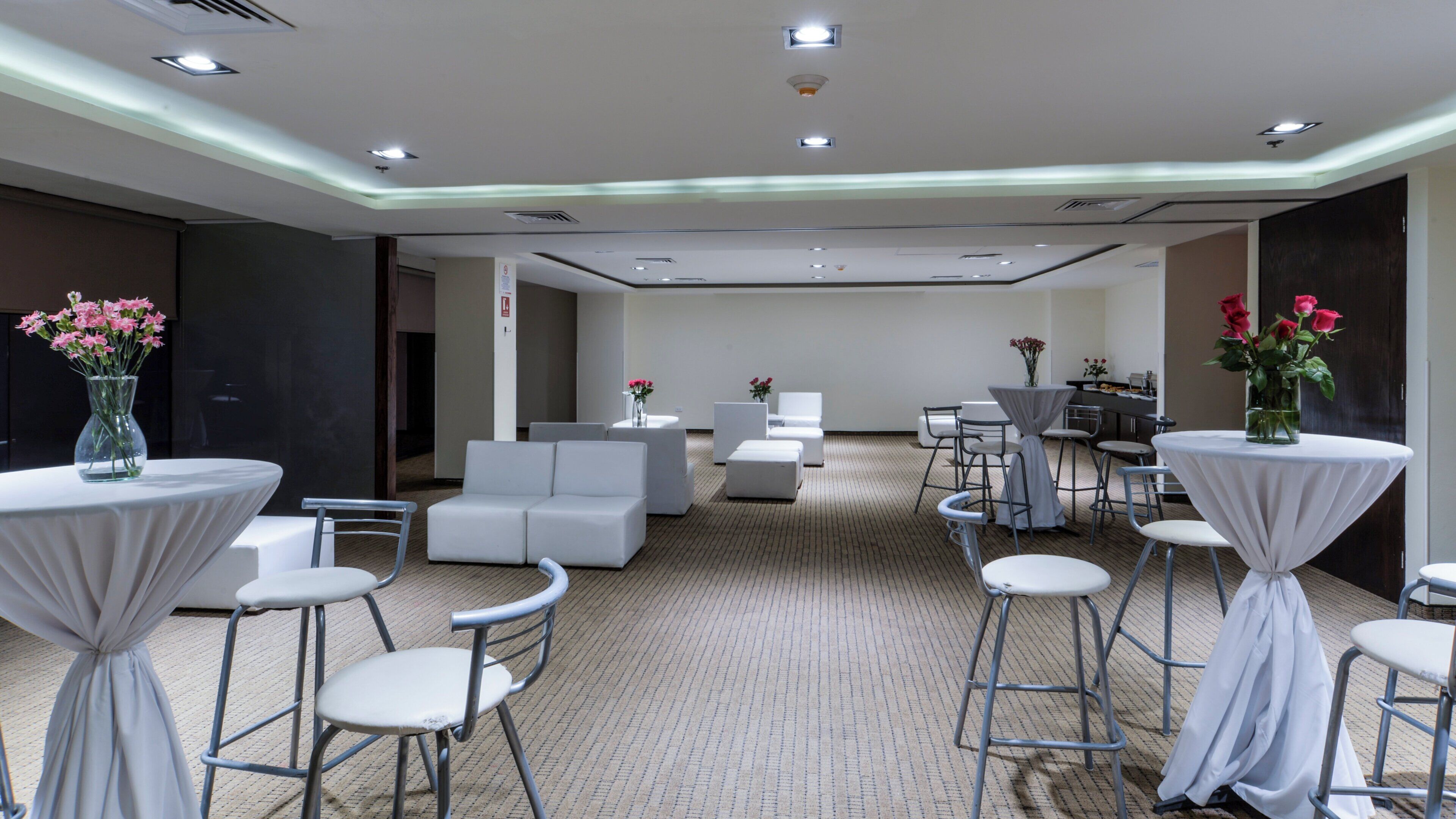Un salón de conferencias en un hotel con mobiliario blanco, mesas con manteles blancos y jarrones de flores.