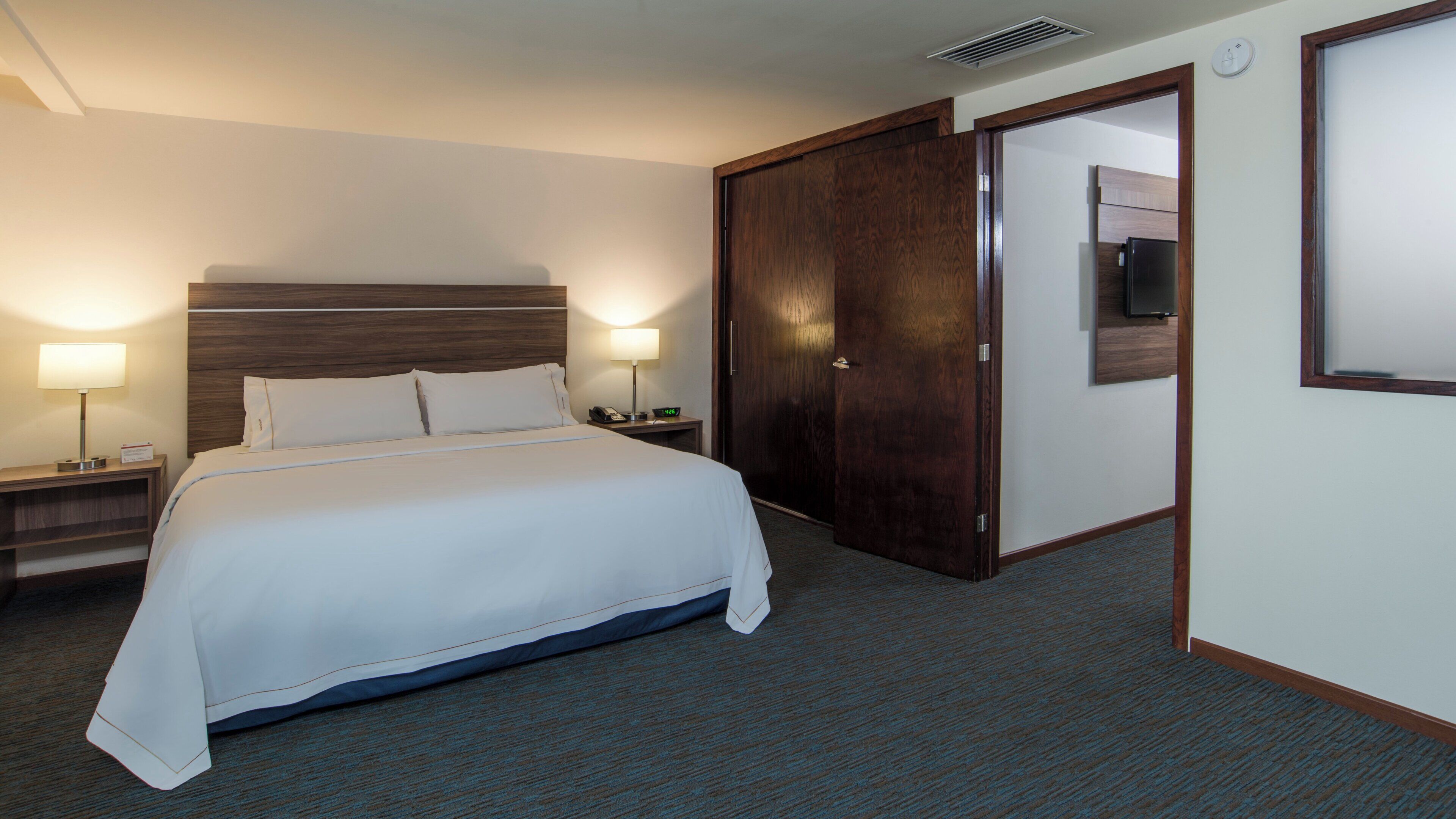 Habitación de hotel con una cama grande, dos mesitas de noche con lámparas, un armario de madera y un televisor en la pared.