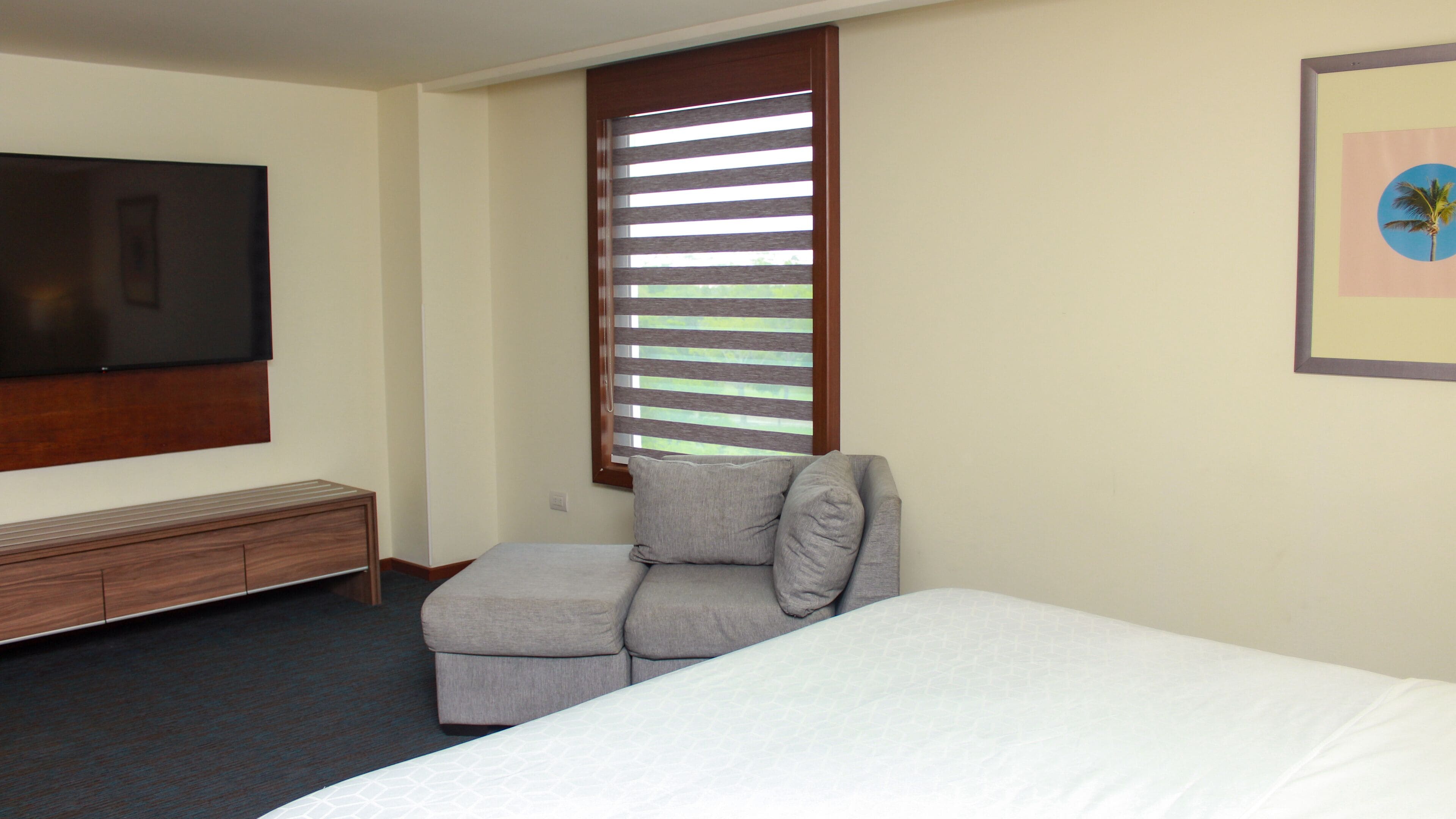 A hotel room with a bed, a gray armchair, a TV mounted on the wall, and a window with blinds.