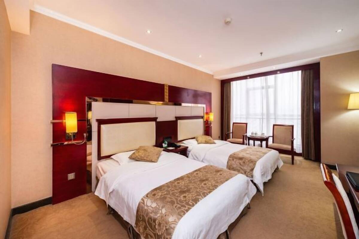 Quarto de hotel com duas camas, uma escrivaninha, uma cadeira e uma janela grande.
