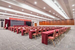 Un salón de conferencias con filas de mesas y sillas, un escenario y suelo alfombrado.