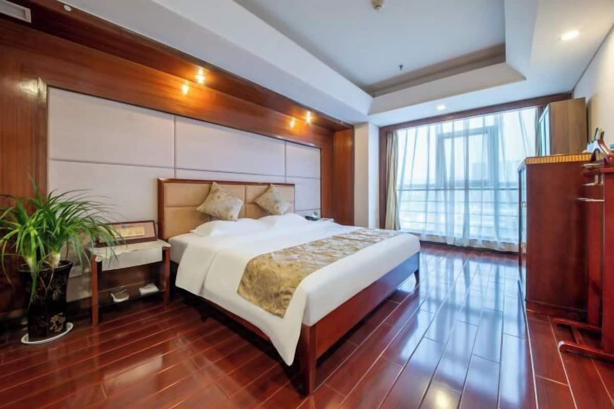 Quarto de hotel com piso de madeira, uma cama com encosto, mesa de cabeceira, planta em vaso e uma janela grande com cortinas.
