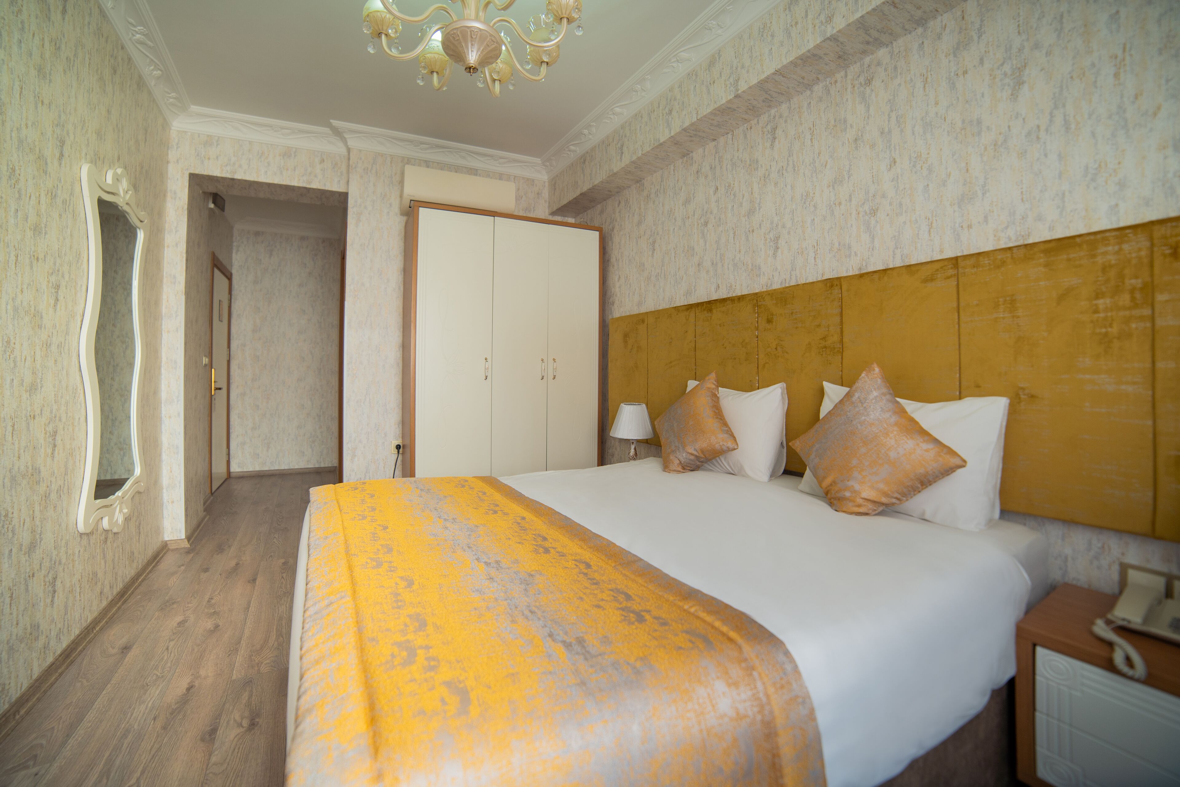 A hotel room with a large bed, a wooden headboard, a mirror, a wardrobe, and a nightstand with a lamp.