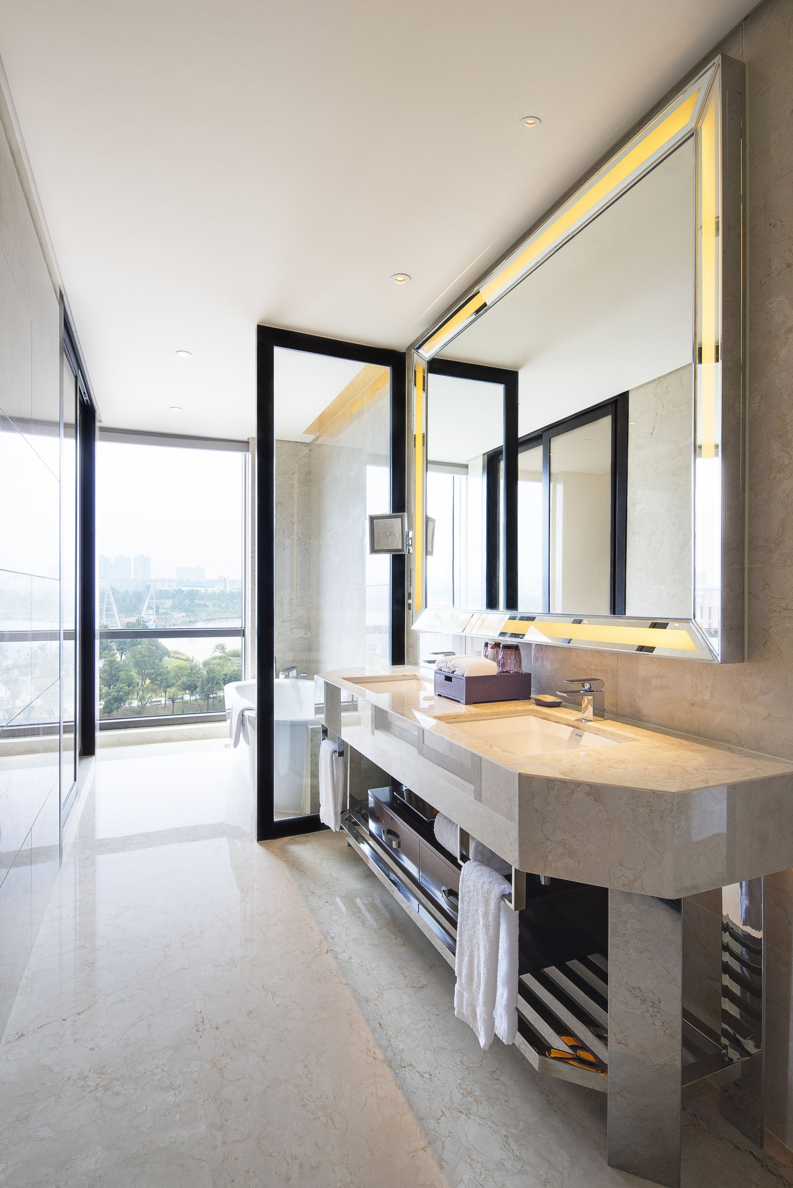 A modern bathroom with a large mirror, dual sinks, and a view of the city through large windows.