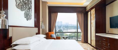A hotel room with a large bed, a TV, a chair, and a window with a city view.