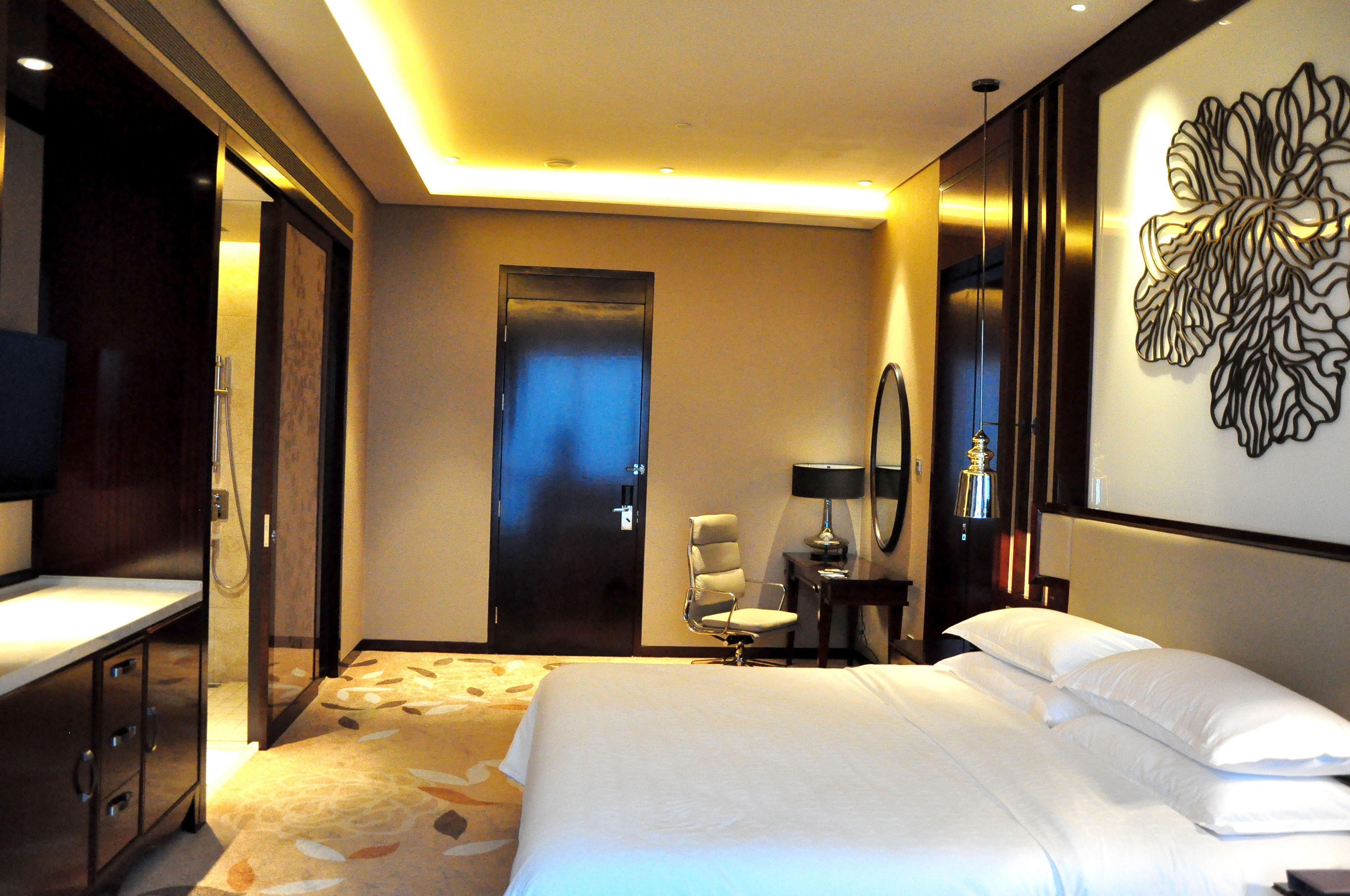 A hotel room with a large bed, a desk with a chair, a mirror, and a wall-mounted artwork.