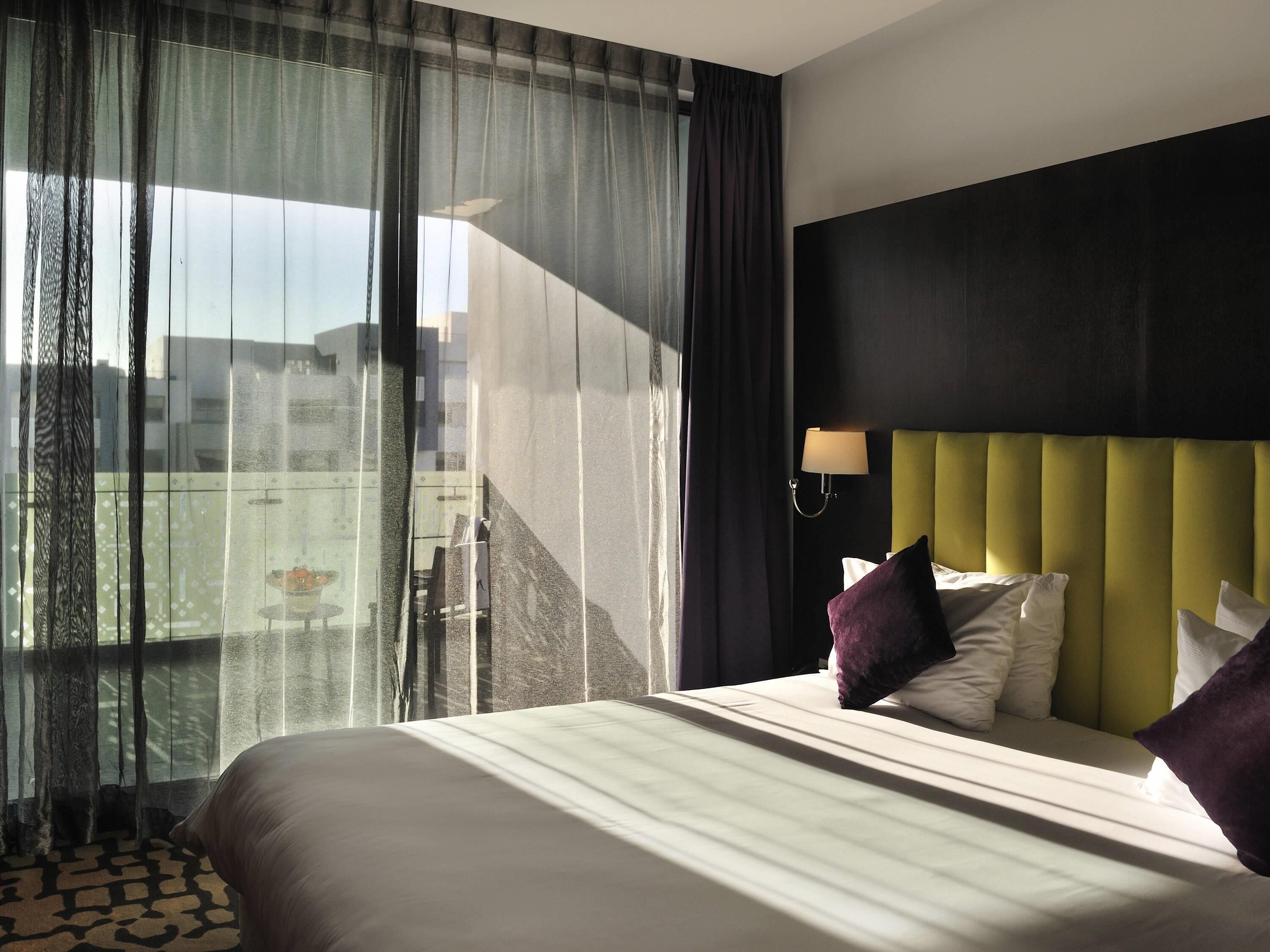 A hotel room with a bed, a headboard, a window with curtains, and a view of the outside.