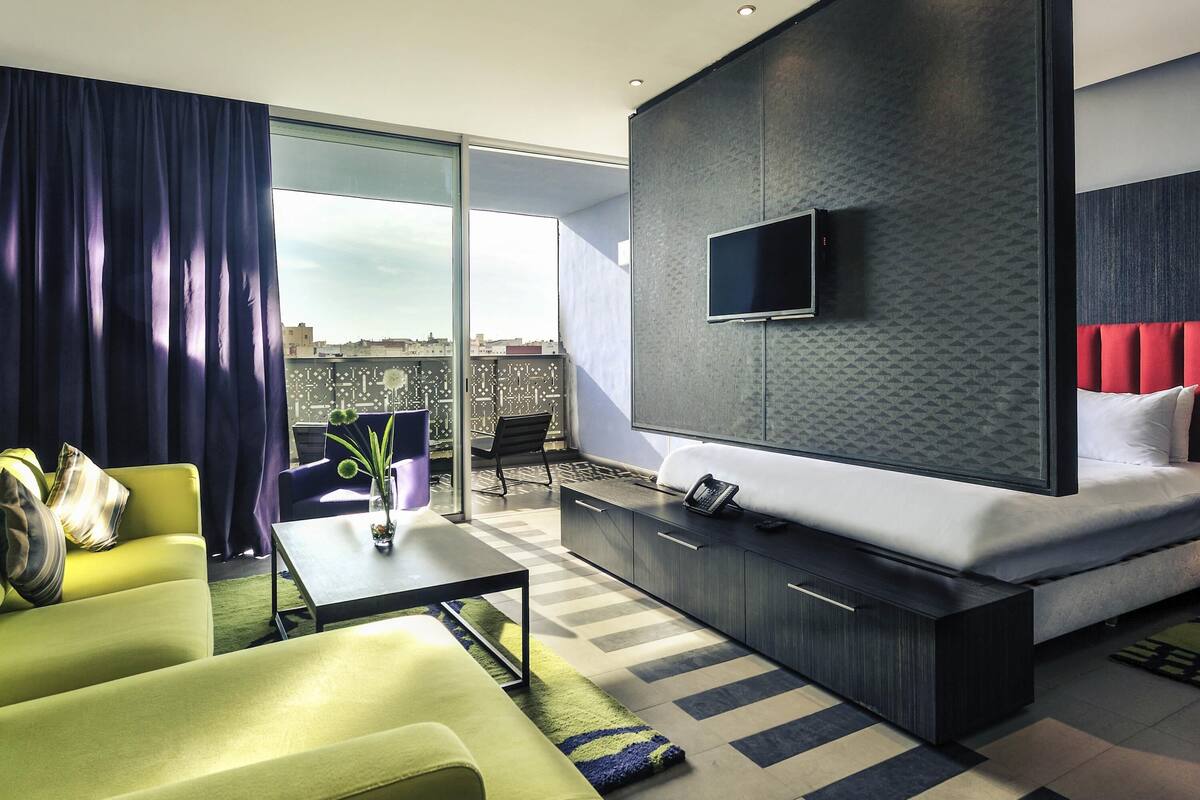 A modern hotel room with a sofa, a small table with a vase, a bed with a red headboard, a TV mounted on the wall, and a balcony with a view.