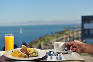 A breakfast setting with a plate of fruit, a glass of orange juice, and a cup, overlooking a scenic view of the sea. - Hotel Mercure Rif Nador (Nador)