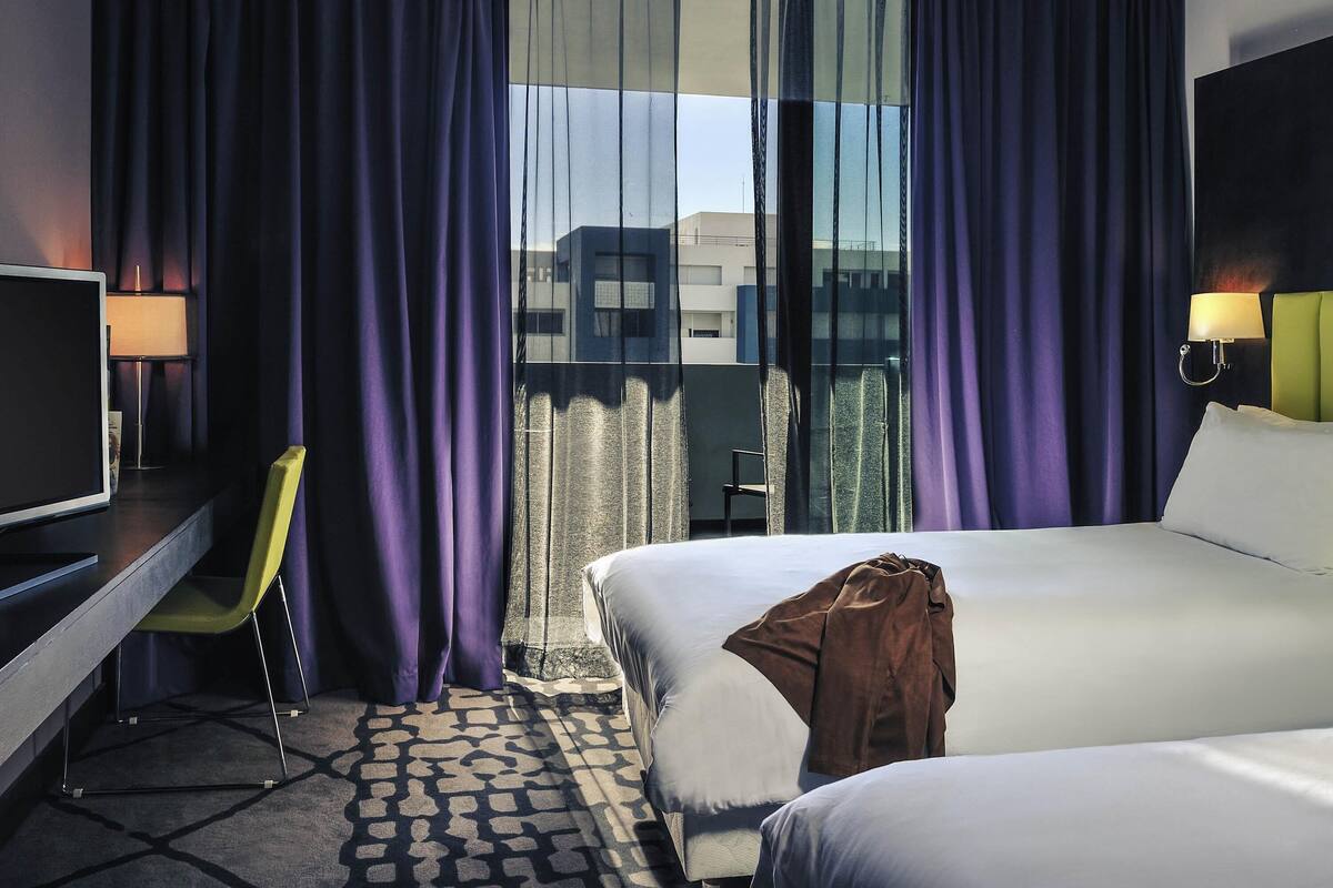 A hotel room with a bed, a desk with a TV, a chair, and a view of buildings through the window.