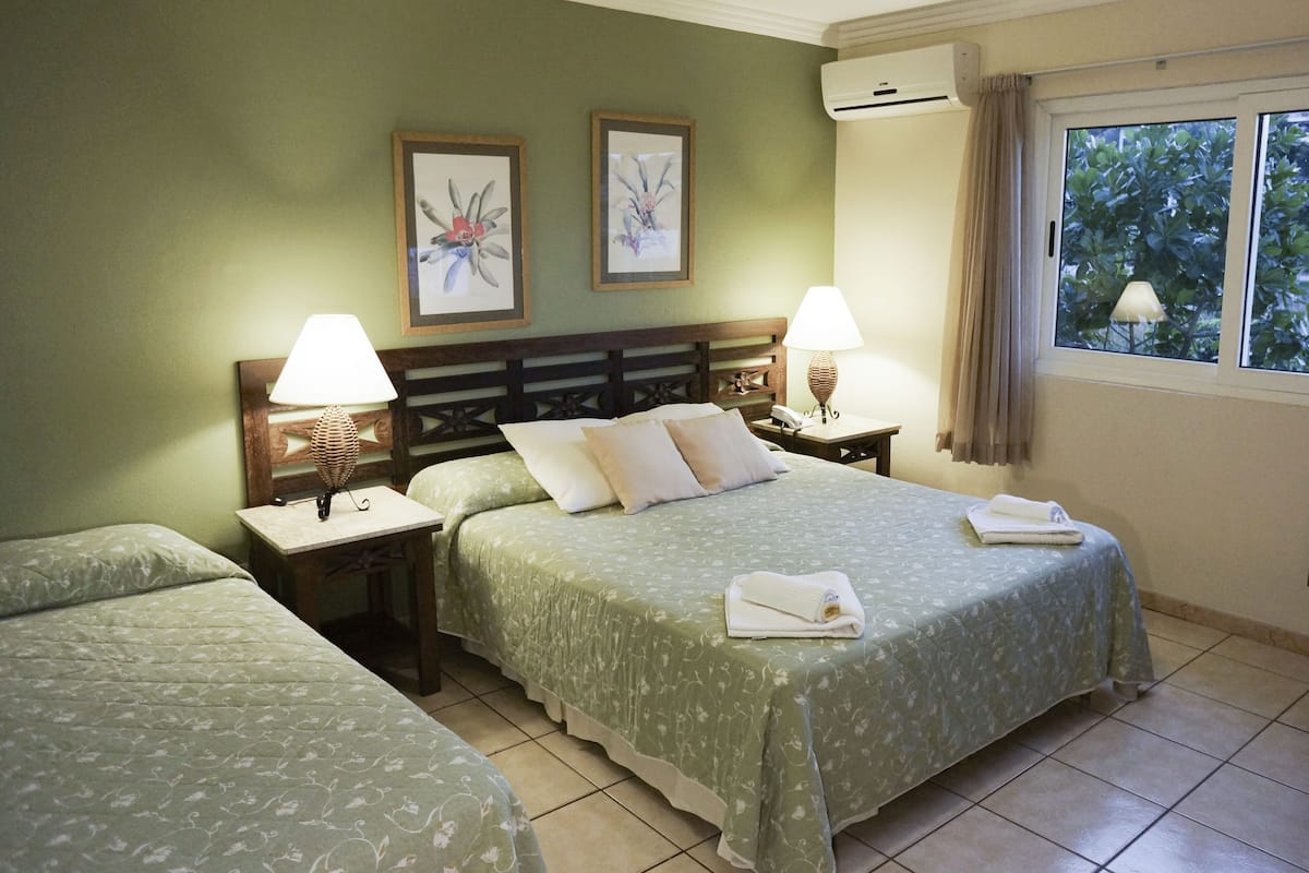 Habitación de hotel con dos camas, cabecera de madera, dos mesitas de noche con lámparas y ventana con vistas a zonas verdes.
