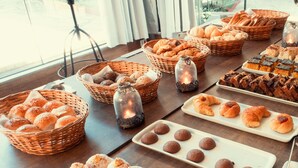 A table with various baked goods, including bread, pastries, and cookies, arranged on a wooden table in a well-lit room with large windows.