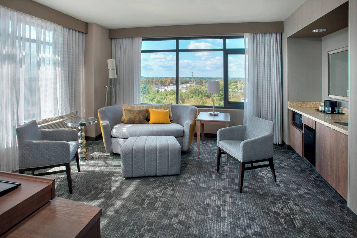 A hotel room with a large window, a sofa, two armchairs, a small table, and a kitchenette.