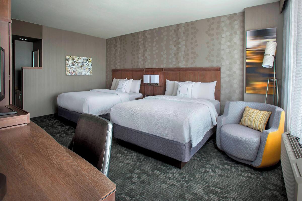 A hotel room with two beds, a desk, a chair, and a large window.