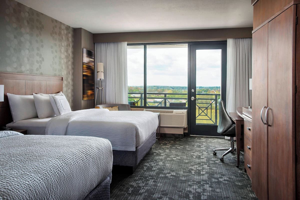A hotel room with two beds, a desk, a chair, and a large window with a balcony view.