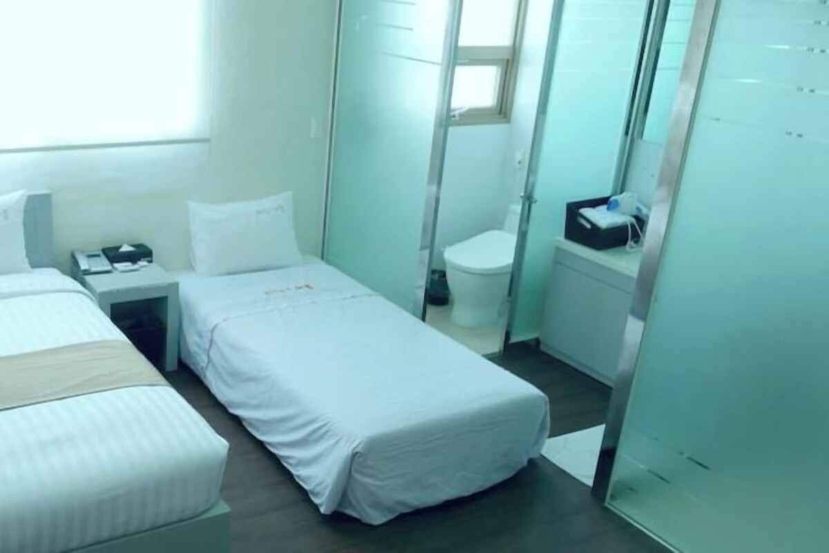 Habitación de hotel con dos camas, una mesita de noche, un escritorio pequeño, una silla, un ventanal y un baño.