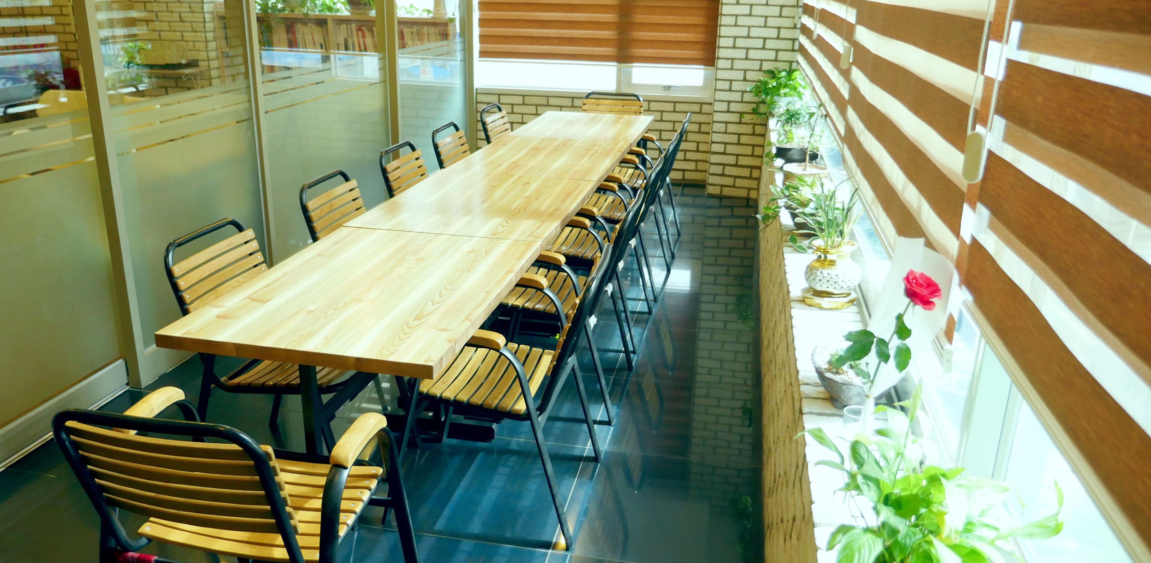 Una larga mesa de conferencias de madera con sillas, rodeada de paredes de vidrio y plantas en macetas.