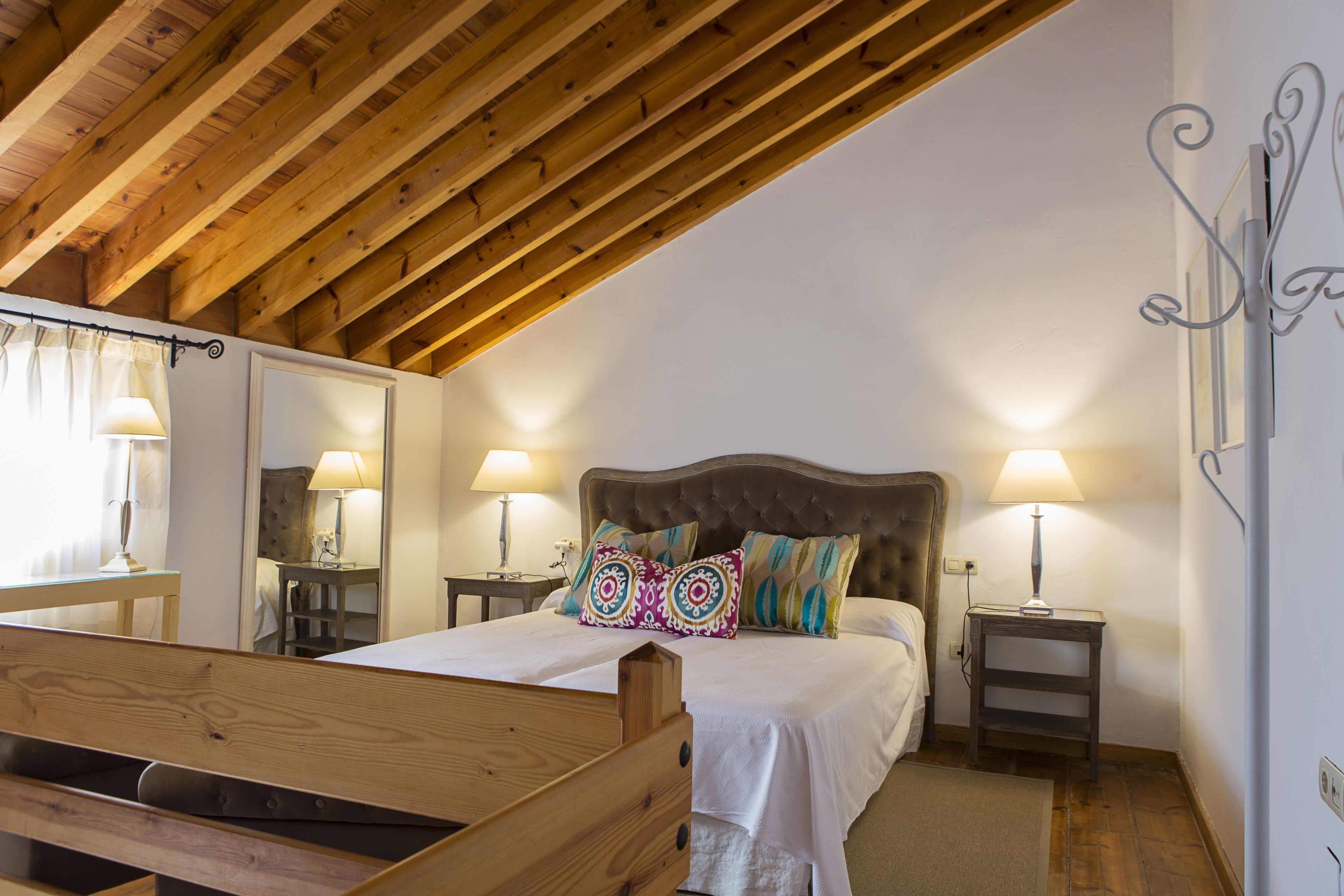 A bedroom with a wooden ceiling, a bed with patterned pillows, bedside lamps, and a wooden bench.
