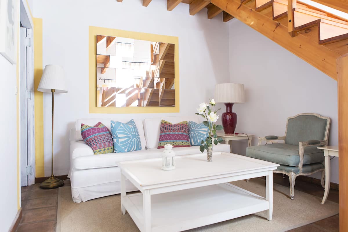 Ein Wohnzimmer mit einem weißen Sofa, bunten Kissen, einem weißen Couchtisch, einer roten Vase, einer Lampe und einem Sessel.