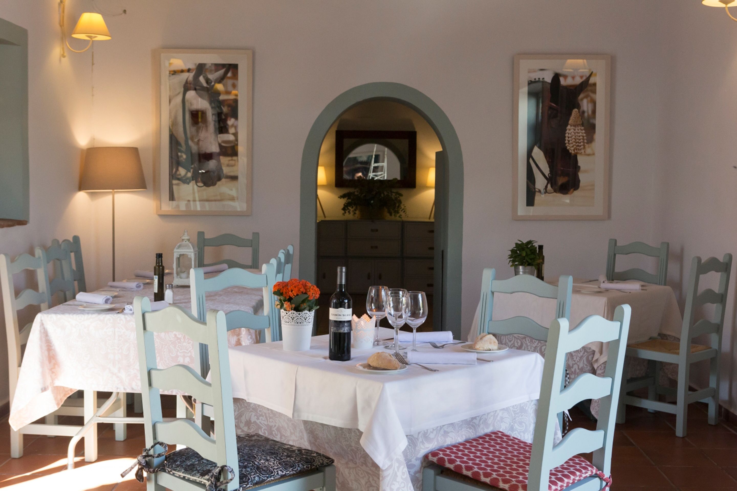 Un comedor con una mesa preparada para dos personas, sillas, una botella de vino, copas y un florero con flores.