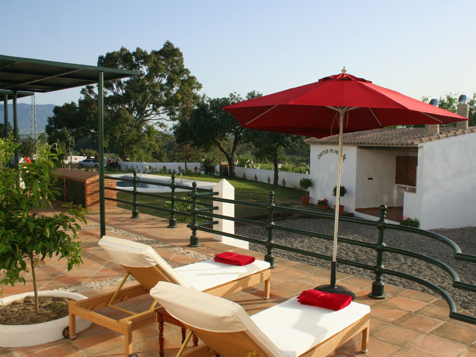 A patio with two sun loungers, a red umbrella, and a small white building labeled "Cortijo del Arco".