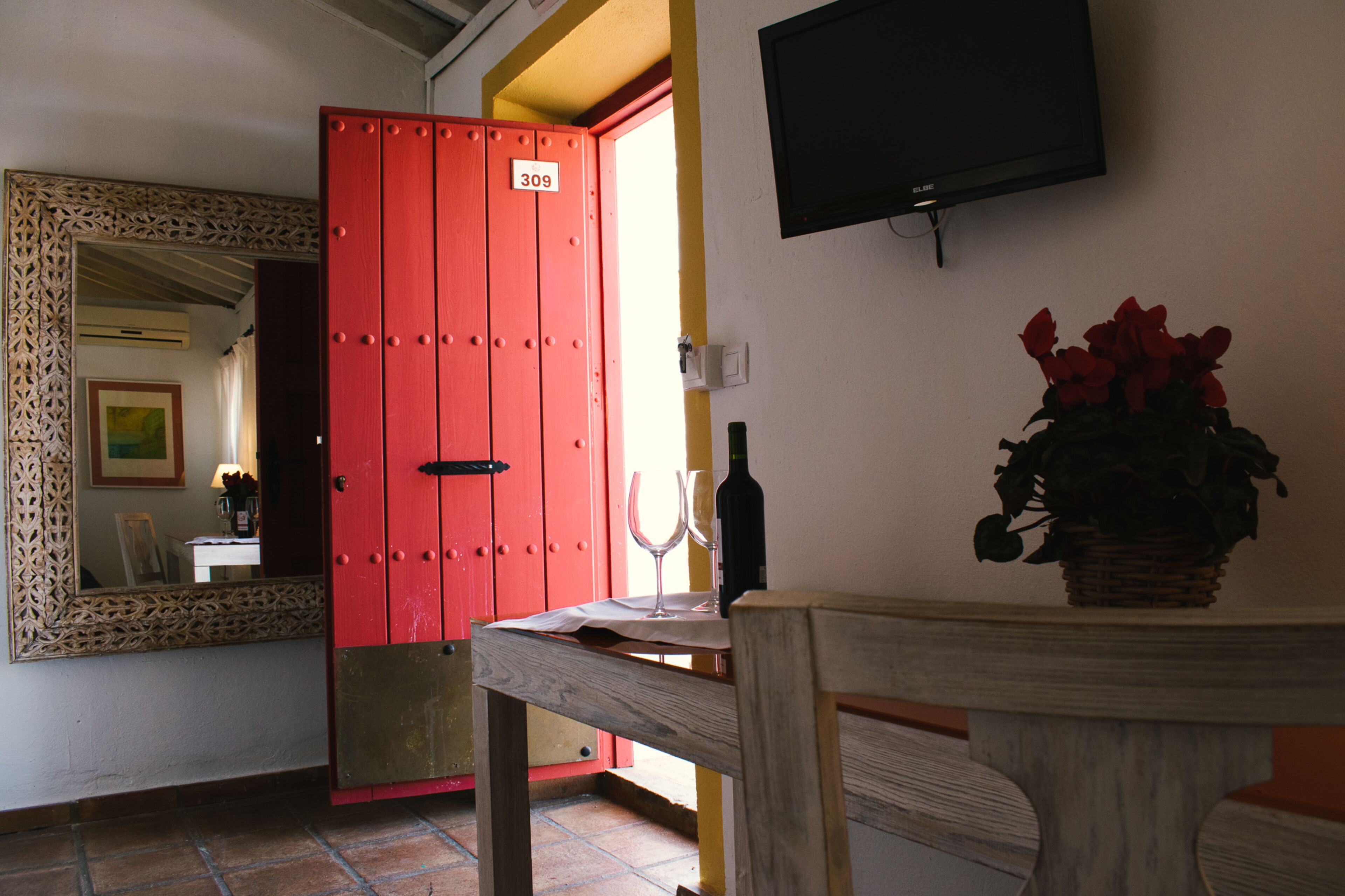 A room with a red door, a wooden table, a bottle, and a vase of flowers.
