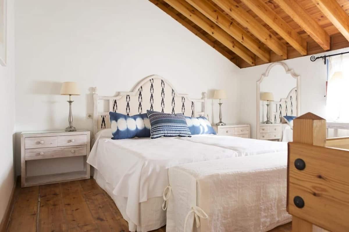 Ein Schlafzimmer mit Holzbalkendecke, einem weißen Bett mit gemusterten Kissen, zwei Nachttischen mit Lampen und einer Holzkommode.