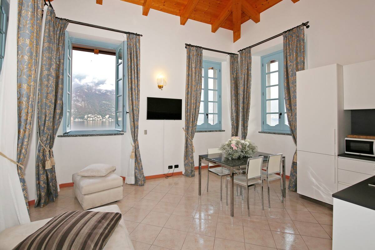 A room with a dining table, chairs, a sofa, a TV mounted on the wall, and a kitchen area.
