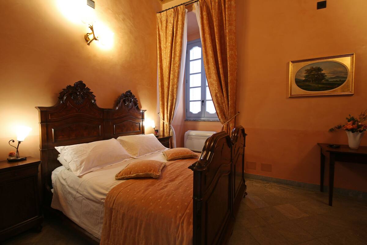 A bedroom with a wooden bed, white bedding, a nightstand with a lamp, a window with curtains, and a framed picture on the wall.