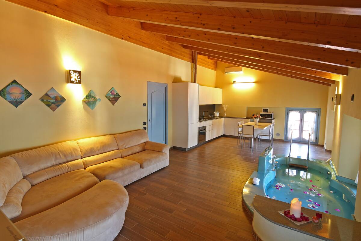 A spacious living area with a wooden ceiling, a large beige sofa, a kitchen area, and a small indoor pool with candles.