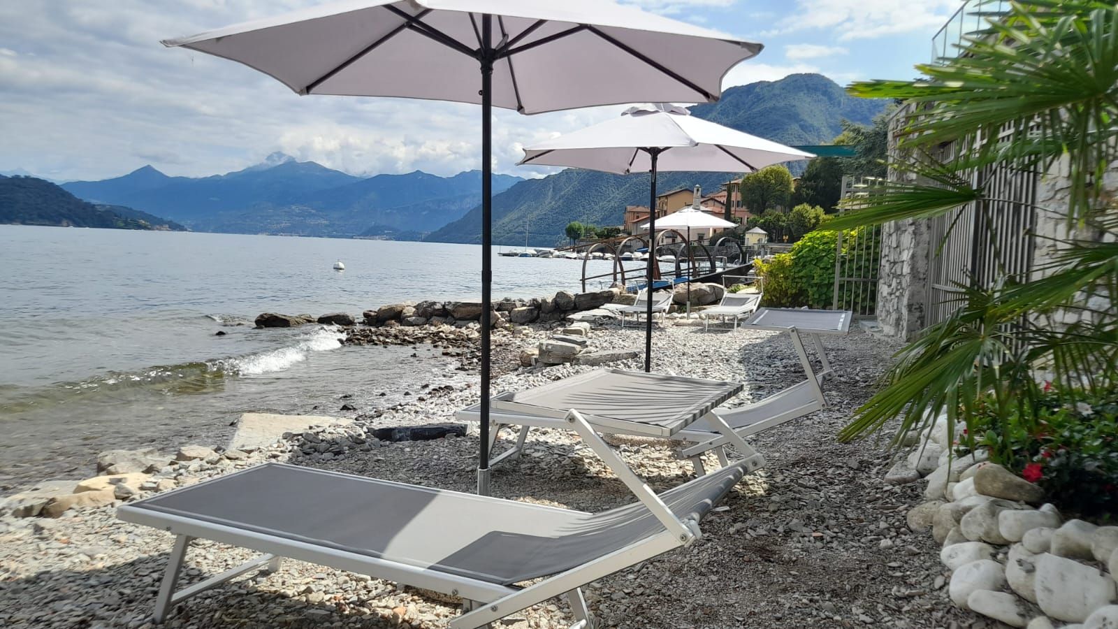 En terrasse ved søen med liggestole og parasoller, en stenet strand og bjerge i baggrunden.