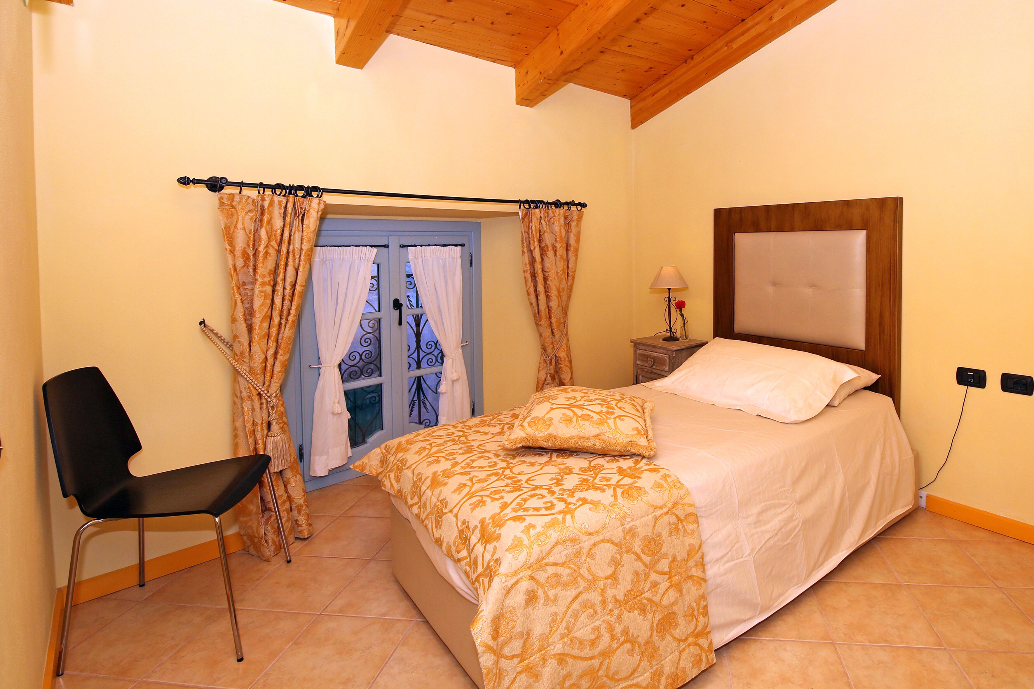 Un dormitorio con cama, una silla, una mesita de noche con lámpara y una ventana con cortinas.