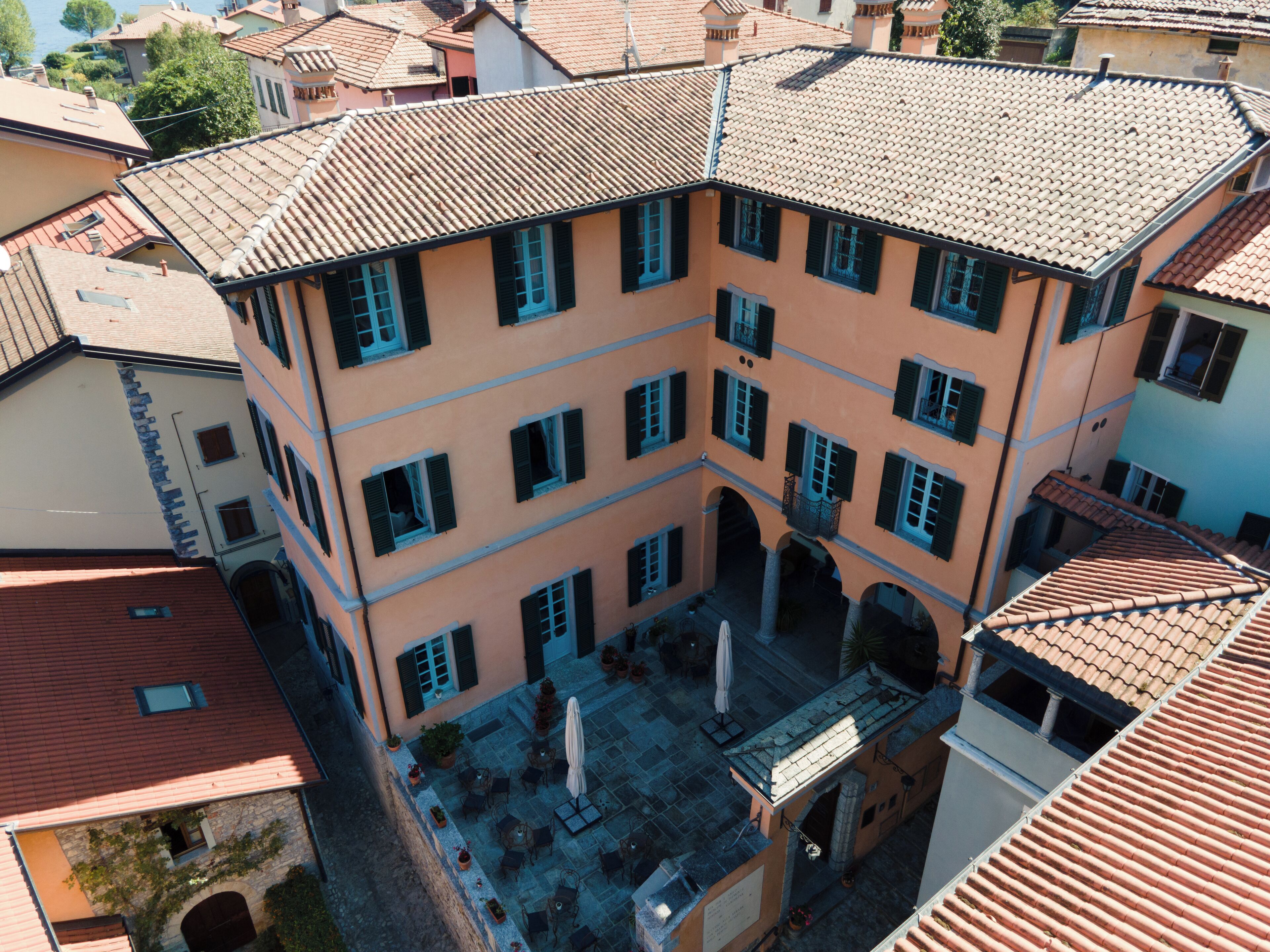 A multi-story building with a courtyard, featuring a tiled roof, multiple windows with shutters, and outdoor seating areas.