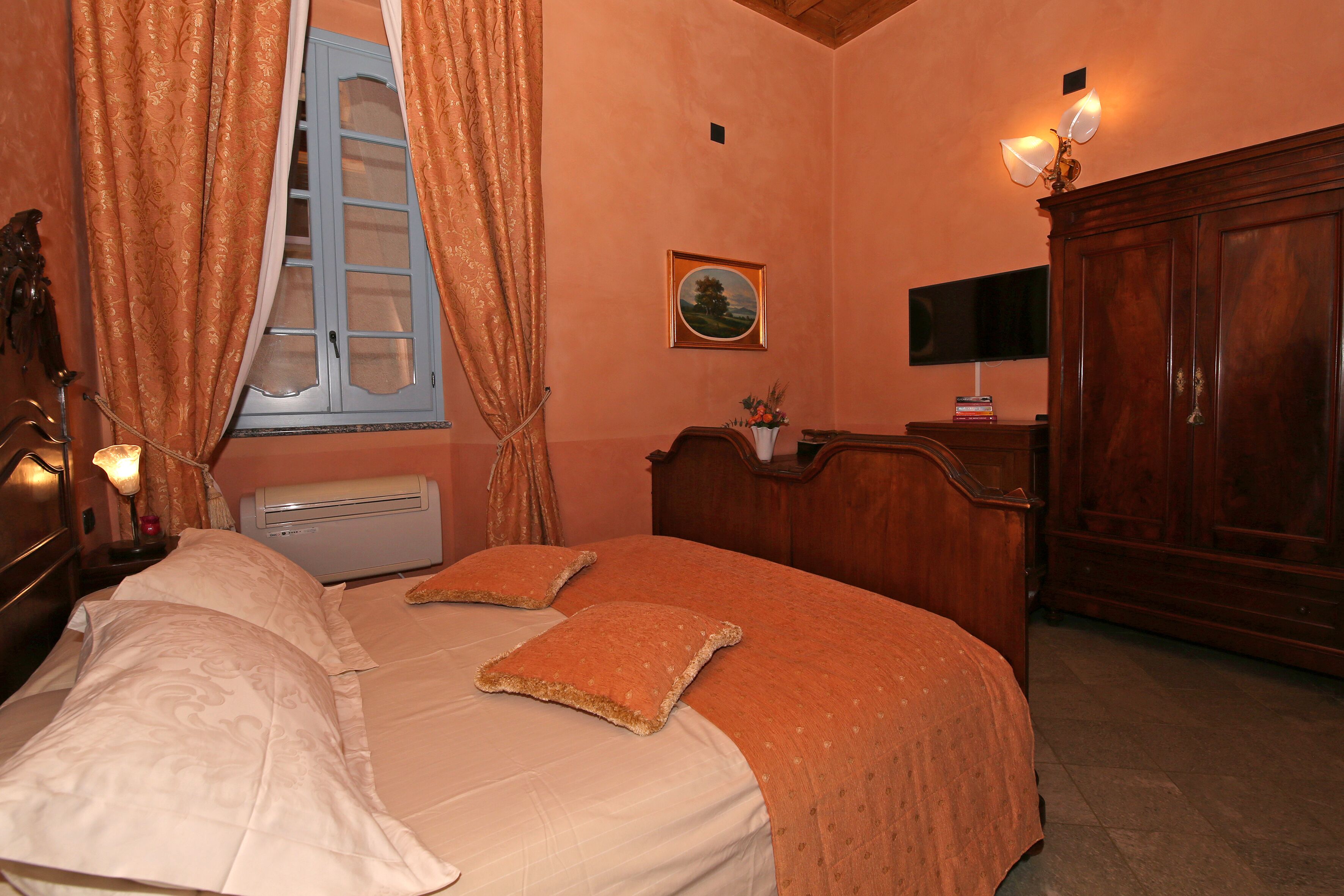 Un dormitorio con cama, cortinas, una mesita de noche, una lámpara, un televisor montado en la pared y un armario de madera.