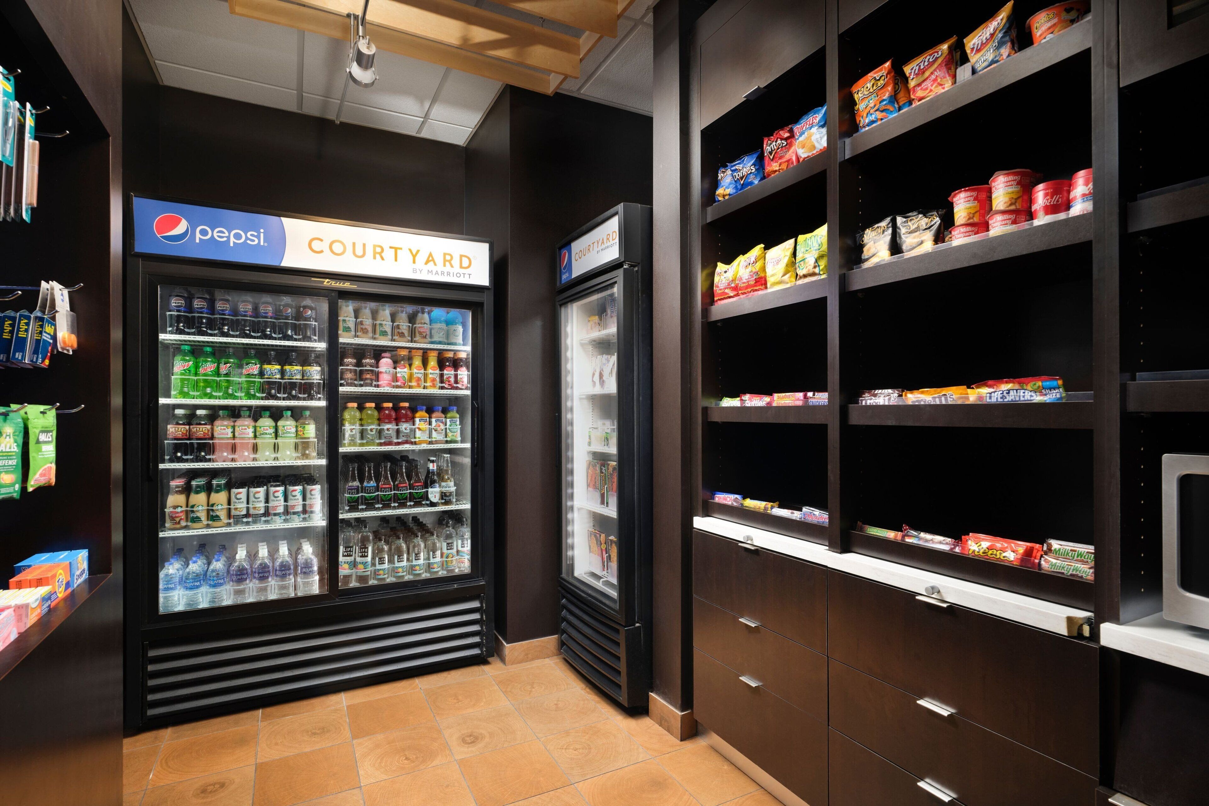 A hotel kitchenette with a Pepsi refrigerator, a microwave, and shelves stocked with snacks and beverages.