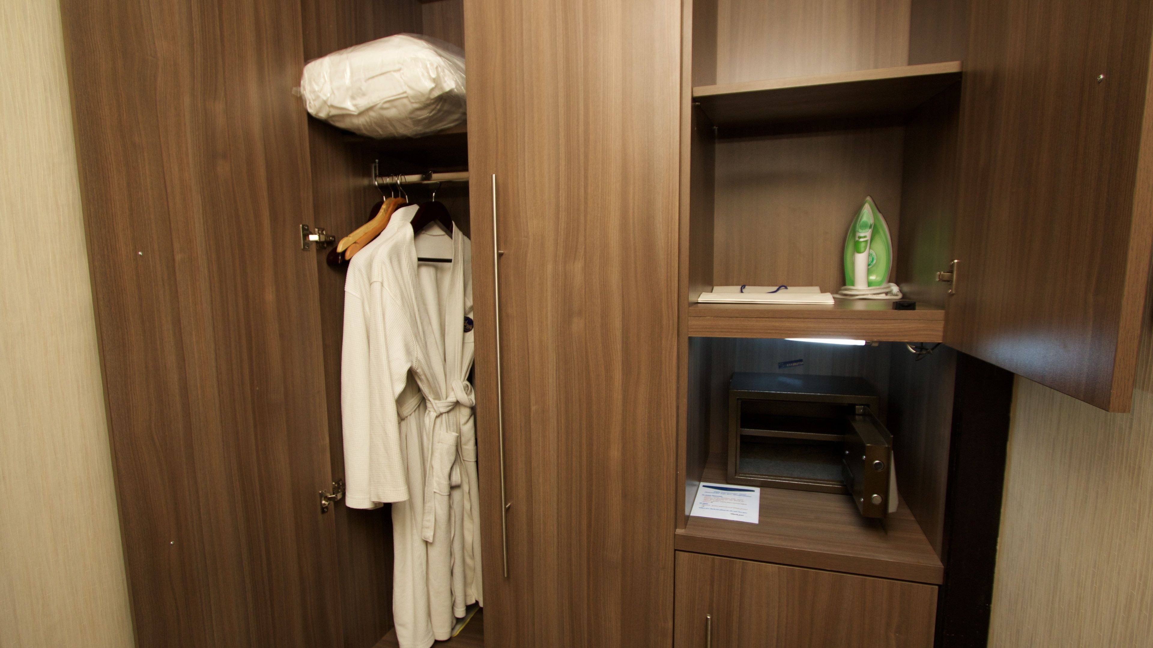 A wooden wardrobe with a hanging robe and a shelf with a green iron and a small item on it.
