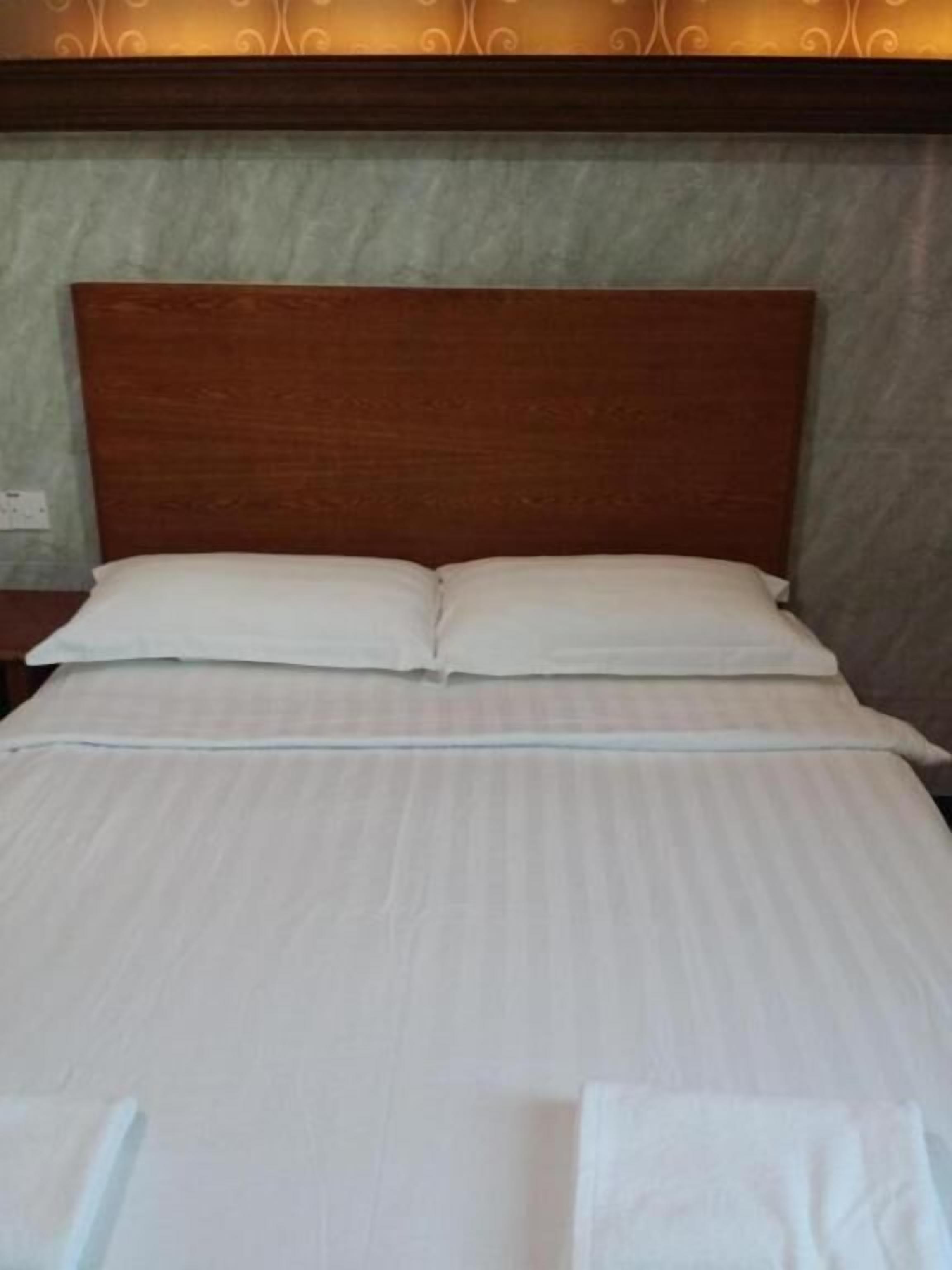 A neatly made bed with a wooden headboard, white linens, and a plain wall with a subtle pattern.