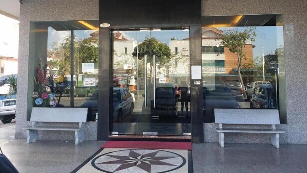 A modern building entrance with a glass facade, a central door, and a red carpet leading to a bench.