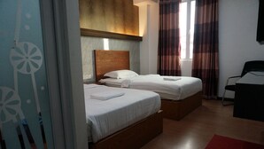 A hotel room with two beds, a wooden headboard, a window with curtains, a desk, and a red rug.