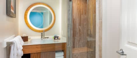 Un baño moderno con ducha de vidrio, tocador de madera y espejo redondo, y toallas ordenadas.