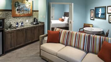 Una habitación de hotel con kitchenette, un sofá con cojines a rayas, una cama con ropa de cama blanca y un área de comedor con mesa y sillas.