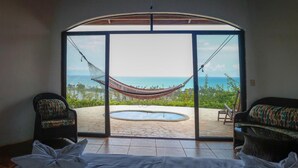 Habitación con un amplio ventanal corredero que da a un patio con una hamaca, una piscina y vista al océano. Hay dos sillas de mimbre y una cama con ropa blanca.