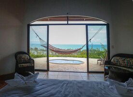 Habitación con un amplio ventanal corredero que da a un patio con una hamaca, una piscina y vista al océano. Hay dos sillas de mimbre y una cama con ropa blanca.