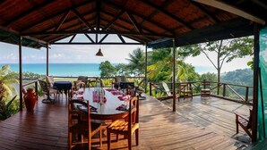 Una terraza de madera con una mesa dispuesta para comer, con vistas a un paisaje tropical y el océano.