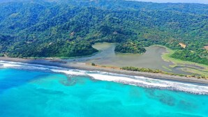 Vista aérea de una zona costera con playa, aguas turquesas cristalinas y una colina boscosa y densa.