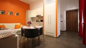 Una sala de estar moderna con sofá, una mesa de comedor con sillas, una zona de cocina con armarios y electrodomésticos, y una puerta de madera.