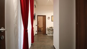 Un pasillo con una puerta a la izquierda, una cortina roja y un estante blanco con objetos a la derecha.