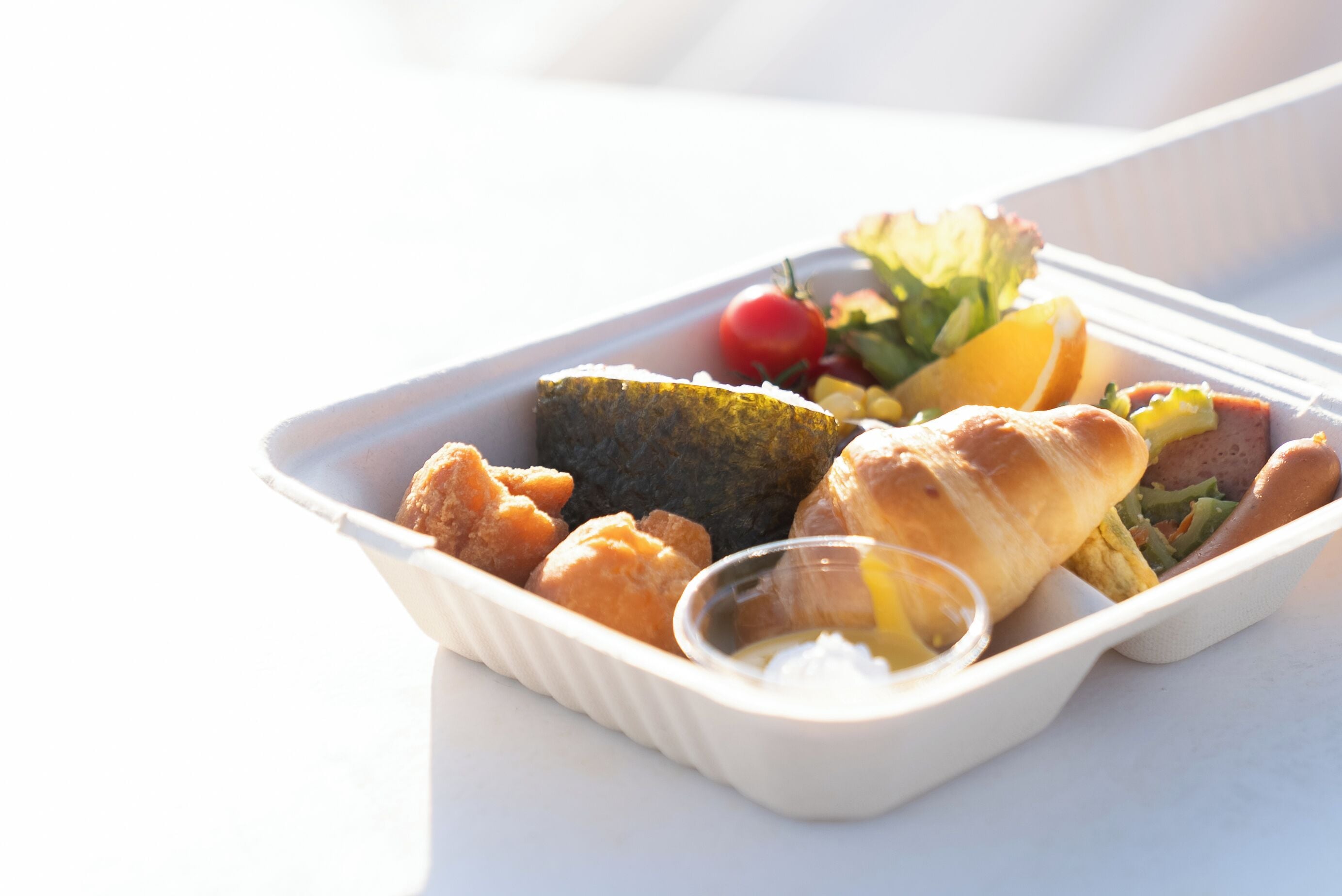 Un repas servi dans une barquette blanche à emporter, contenant divers aliments, notamment du pain, des légumes et une petite portion de sauce.