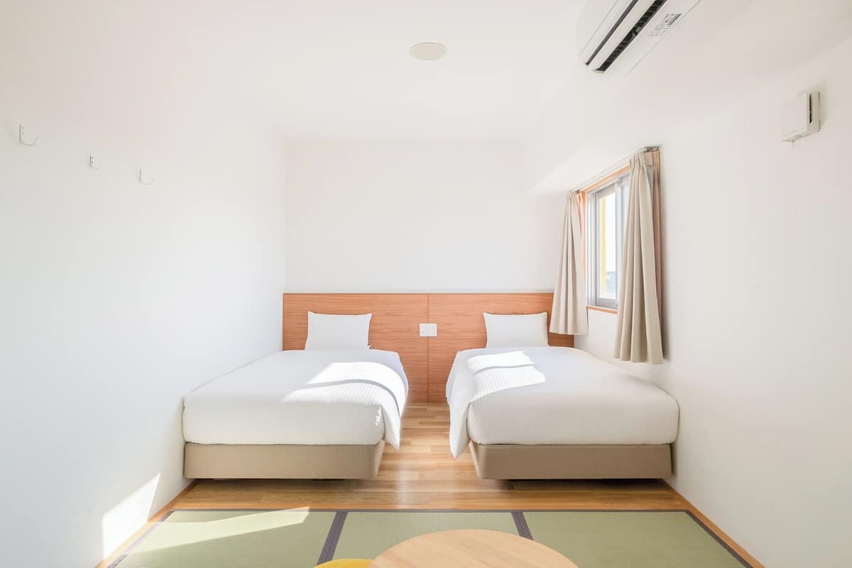 Une chambre avec deux lits, une tête de lit en bois, une fenêtre avec des rideaux et un appareil de climatisation.