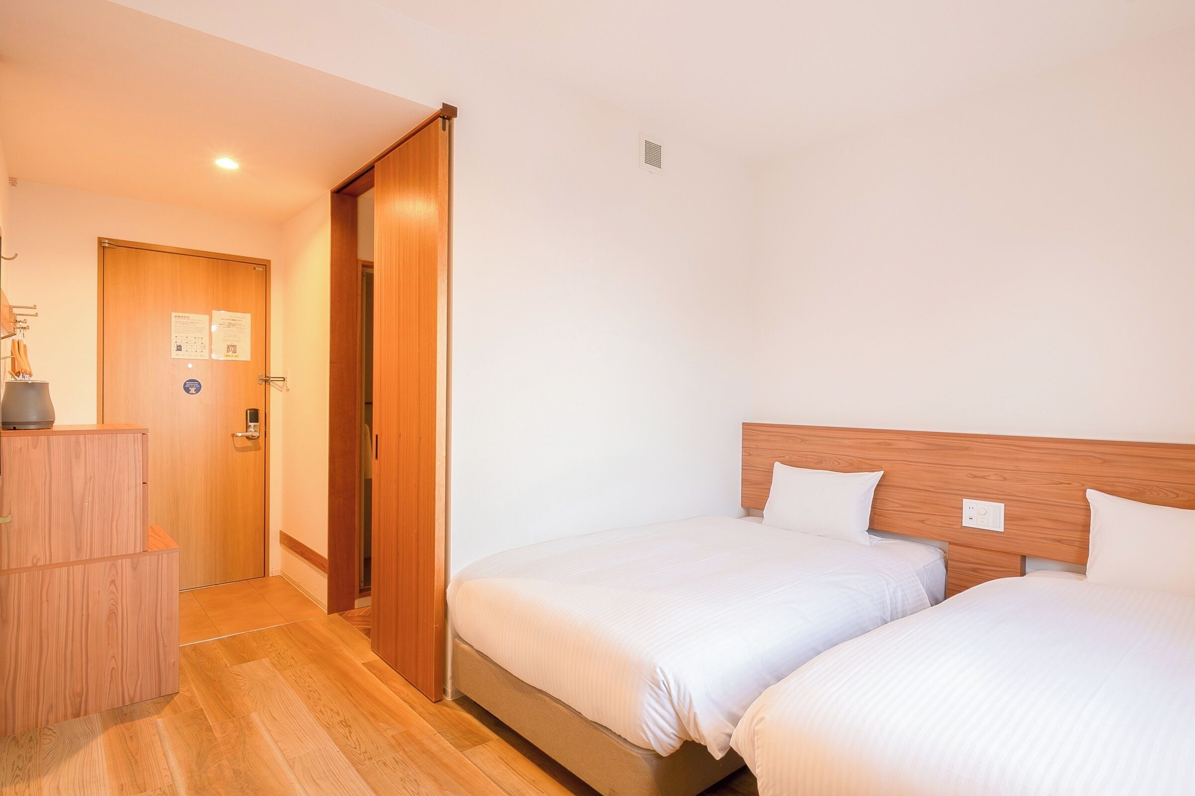 A hotel room with two beds, wooden headboards, a wooden wardrobe, and a door with a notice.